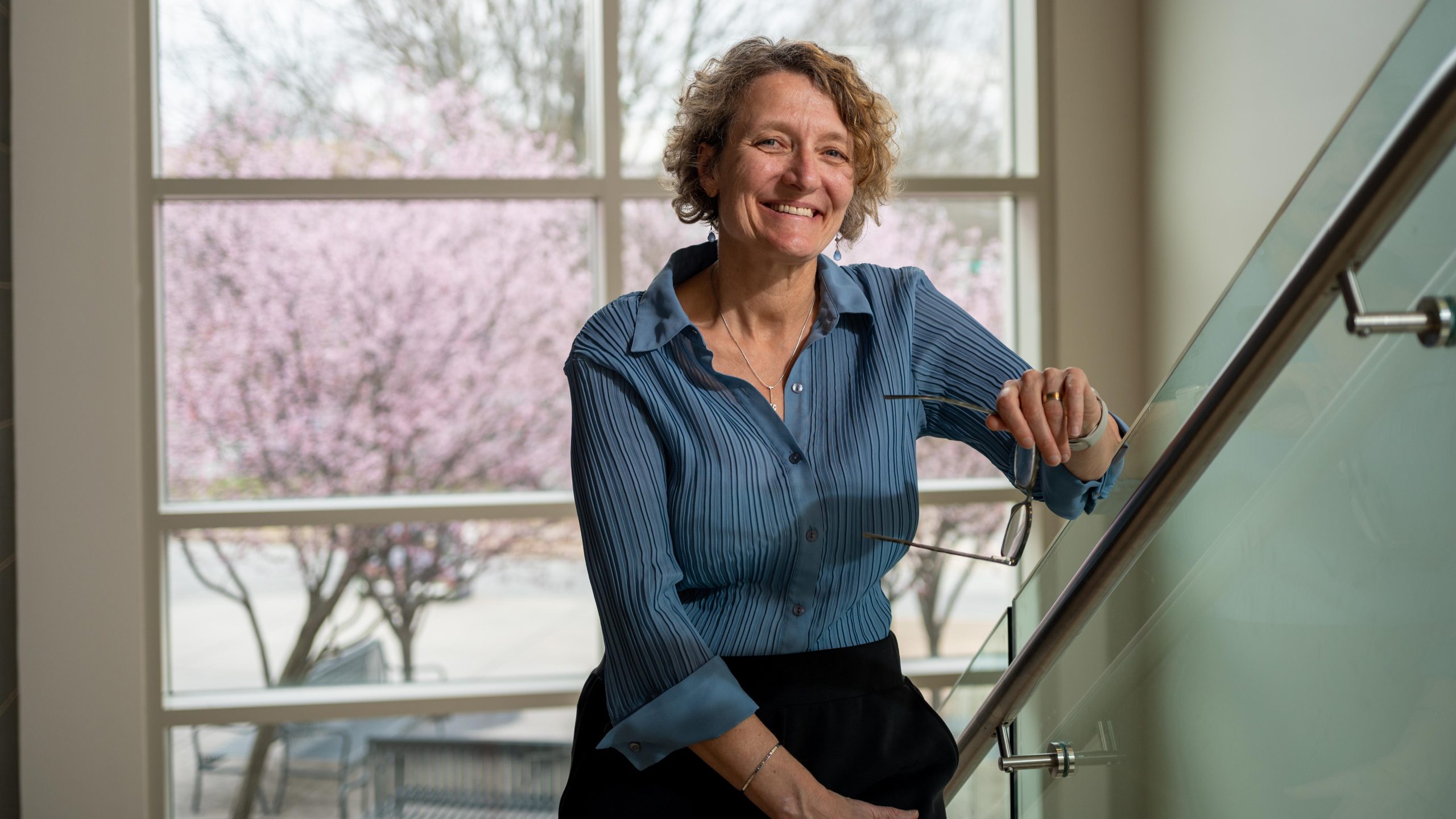 Portrait of UNCG's Julie Edmunds smiling, leaned on staircase holding a pair of glasses